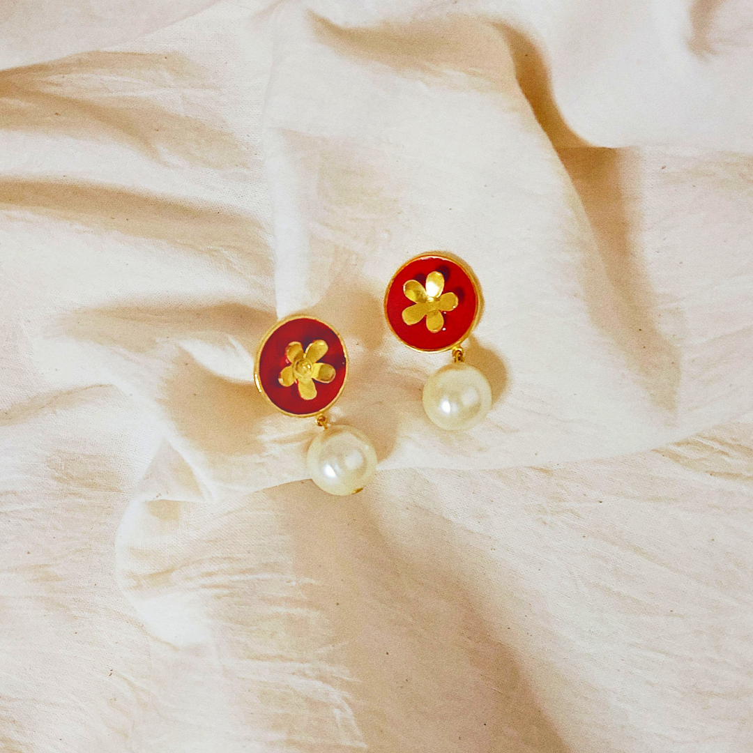 A pair of handcrafted earrings featuring red Meena enamel discs with gold floral motifs and elegant faux pearl drops, set in 18K gold-plated brass.