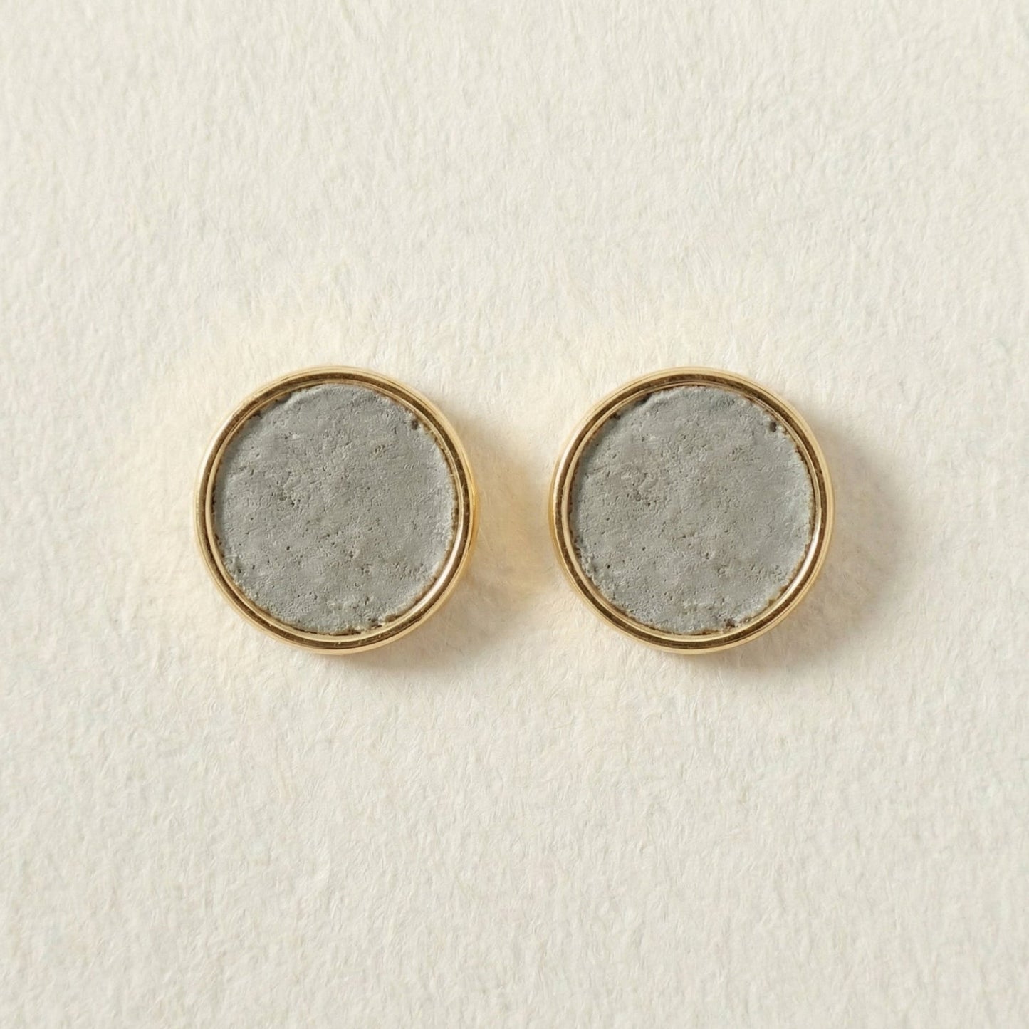 Round cement earrings with gold rims and concrete-like centers on a light beige background
