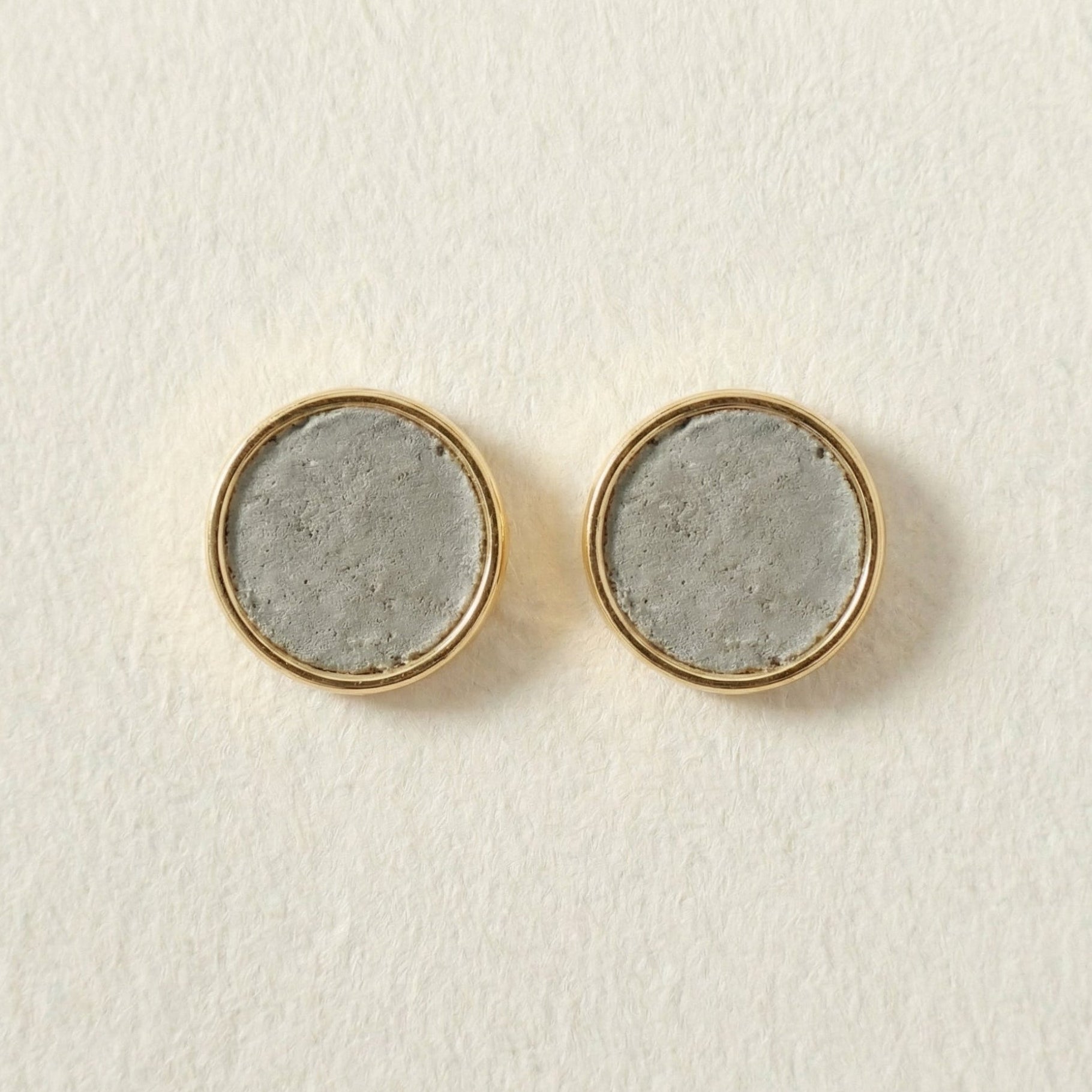 Round cement earrings with gold rims and concrete-like centers on a light beige background